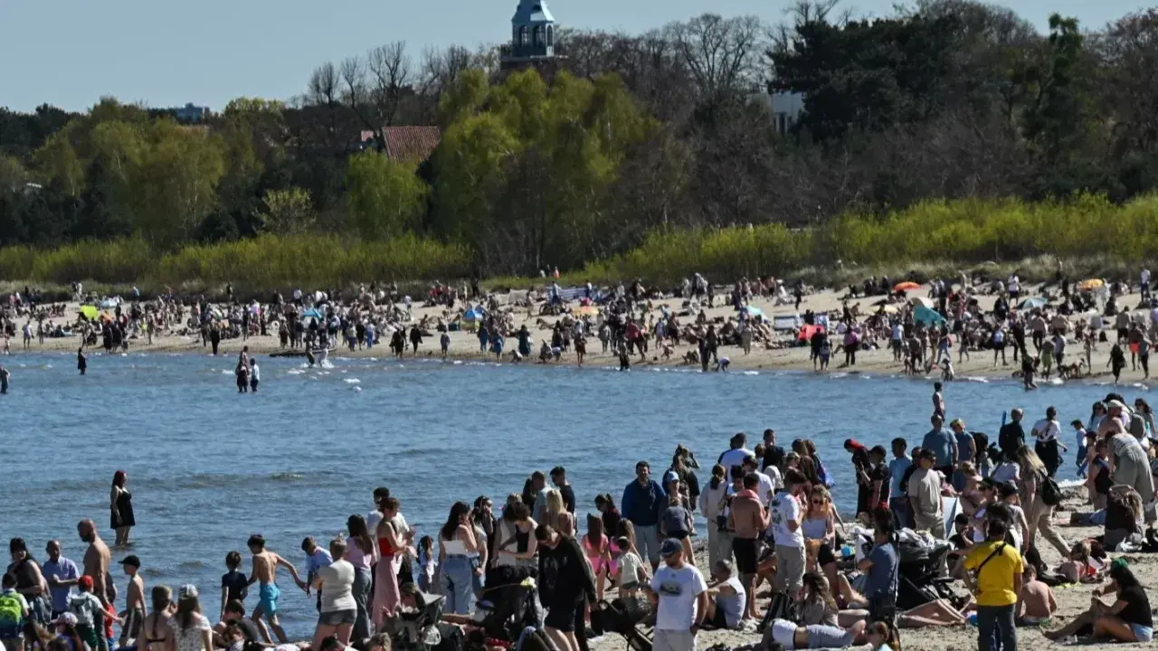 Tłumy ludzi na plaży, woda i zielone drzewa w tle. Widać wieżę kościelną. Ceny nad morzem pewnie wysokie, ale widok piękny.