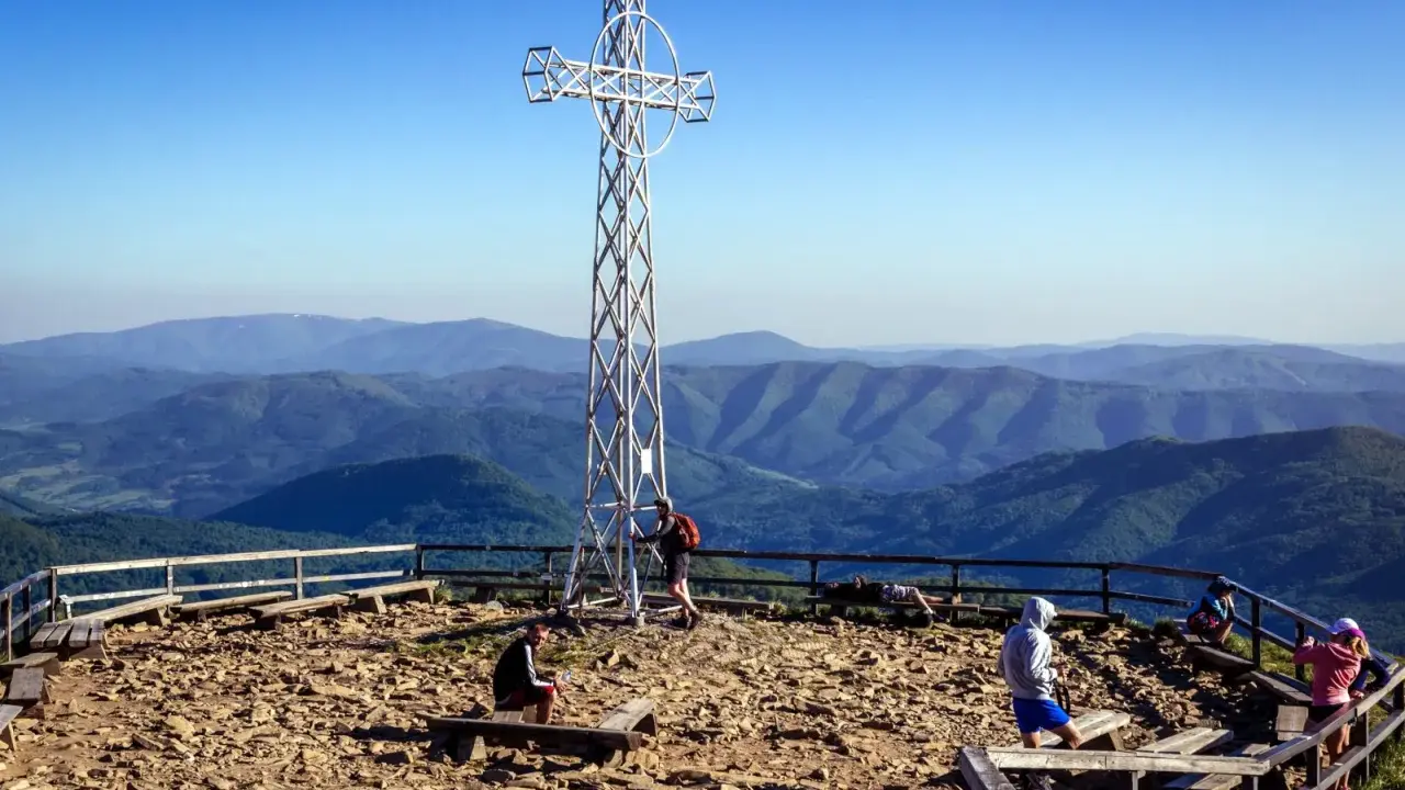 Widok na Bieszczady z krzyżem na szczycie. Ludzie odpoczywają na ławkach, podziwiając rozległe pasma górskie.