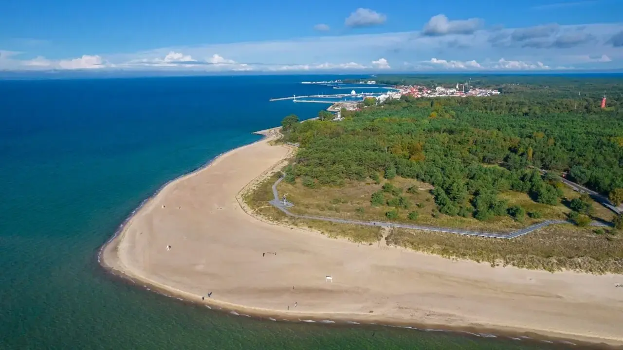 Piękny widok na plażę i las na Helu. Morze o turkusowym odcieniu, piaszczysta plaża z kilkoma spacerującymi osobami, a w oddali widać zabudowania i port.