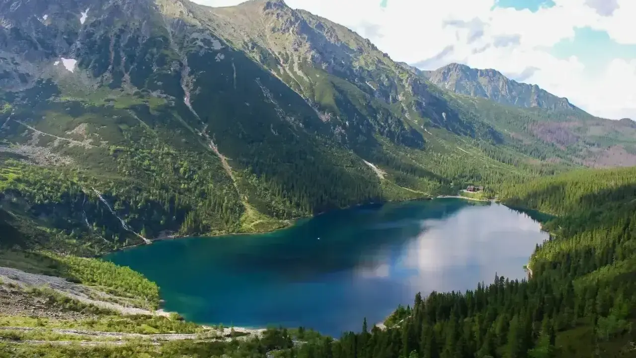 Górskie jezioro otoczone lasem, z wodą odbijającą niebo. W oddali widać skaliste szczyty i łysą polanę.