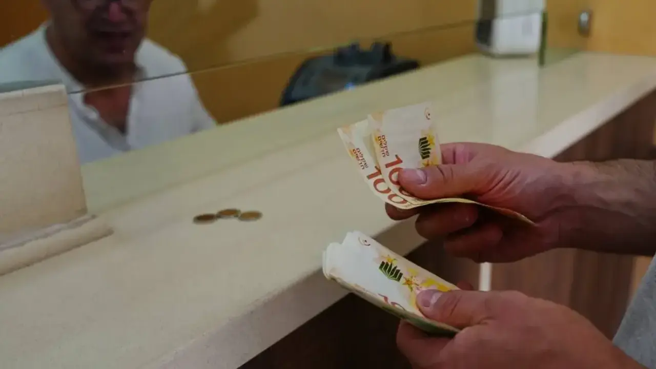 Ręce trzymają banknoty 100 szekli, waluta Izraela. W tle mężczyzna za ladą.