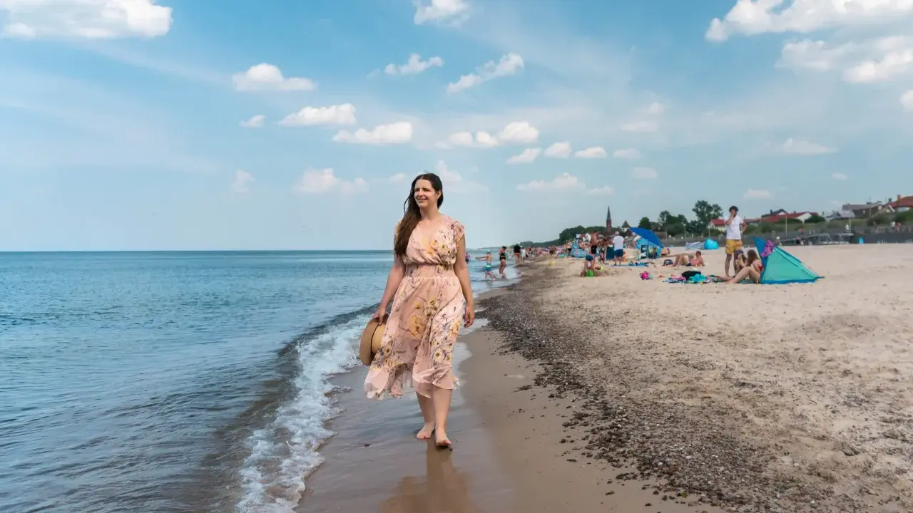 Kobieta w kwiecistej sukience spaceruje brzegiem morza w Sarbinowie. Na plaży widać ludzi odpoczywających na kocach i pod parasolami.