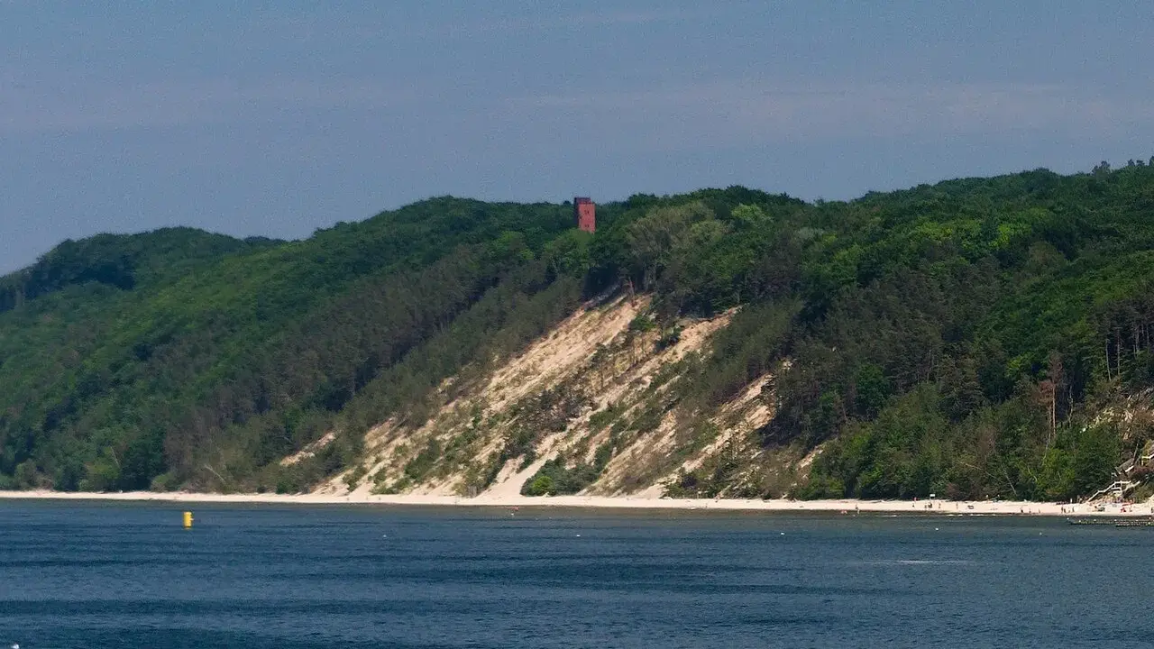 Zielone wzgórza porośnięte lasem schodzą ku piaszczystej plaży. Na szczycie widać czerwoną wieżę. To piękna, biała góra nad morzem.