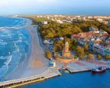 Malownicze miasta nad morzem z latarnią morską, plażą i molo. Widać port z żaglowcem i falujące morze.