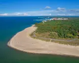 Piękny widok na plażę i las na Helu. Morze o turkusowym odcieniu, piaszczysta plaża z kilkoma spacerującymi osobami, a w oddali widać zabudowania i port.