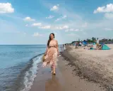 Kobieta w kwiecistej sukience spaceruje brzegiem morza w Sarbinowie. Na plaży widać ludzi odpoczywających na kocach i pod parasolami.