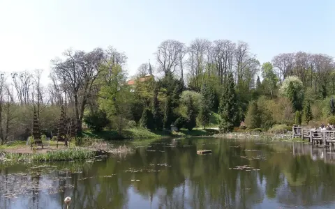 Spokojny staw w Bolestraszycach, otoczony drzewami i zielenią. Widoczna drewniana kładka z ludźmi, idealne miejsce na relaks i podziwianie atrakcji.