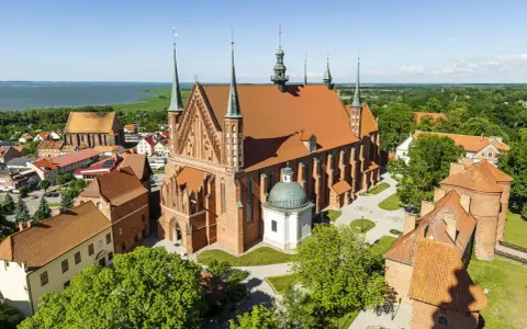 Zamek Frombork z katedrą i wieżami, otoczony zielenią, nad błękitną wodą.