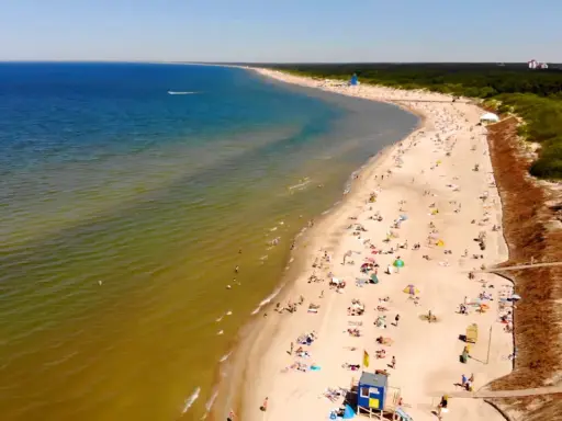 Słoneczna plaża na Litwie, gdzie turkusowe morze łagodnie przechodzi w zielonkawe wody. Tłumy plażowiczów odpoczywają na piasku, ciesząc się letnim dniem.