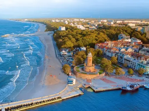 Malownicze miasta nad morzem z latarnią morską, plażą i molo. Widać port z żaglowcem i falujące morze.