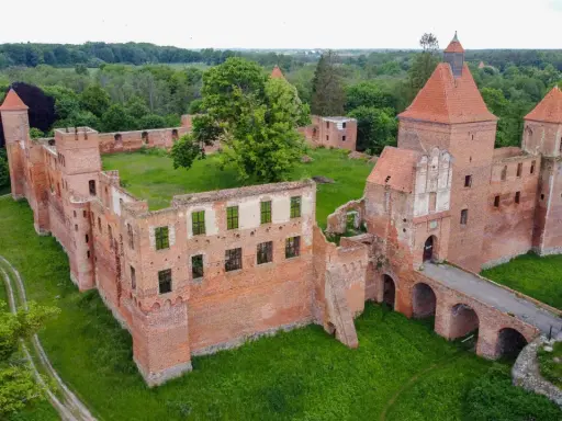 Ruiny ceglanego zamku w Szembarku otoczone zielenią. Widać most nad fosą, wieże i fragmenty murów obronnych.