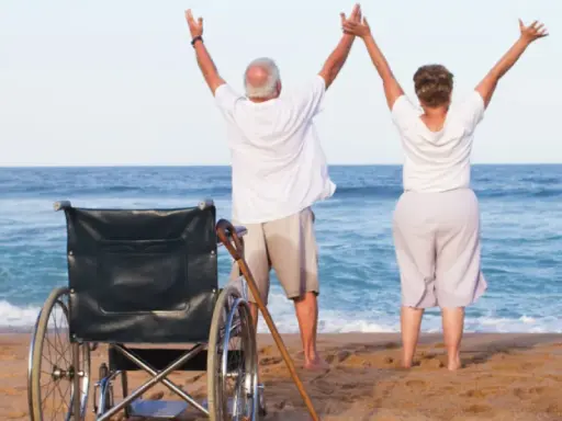Szczęśliwi seniorzy cieszą się urlopem. Wózek inwalidzki i laska na plaży świadczą o tym, że to udany turnus rehabilitacyjny nad morzem.