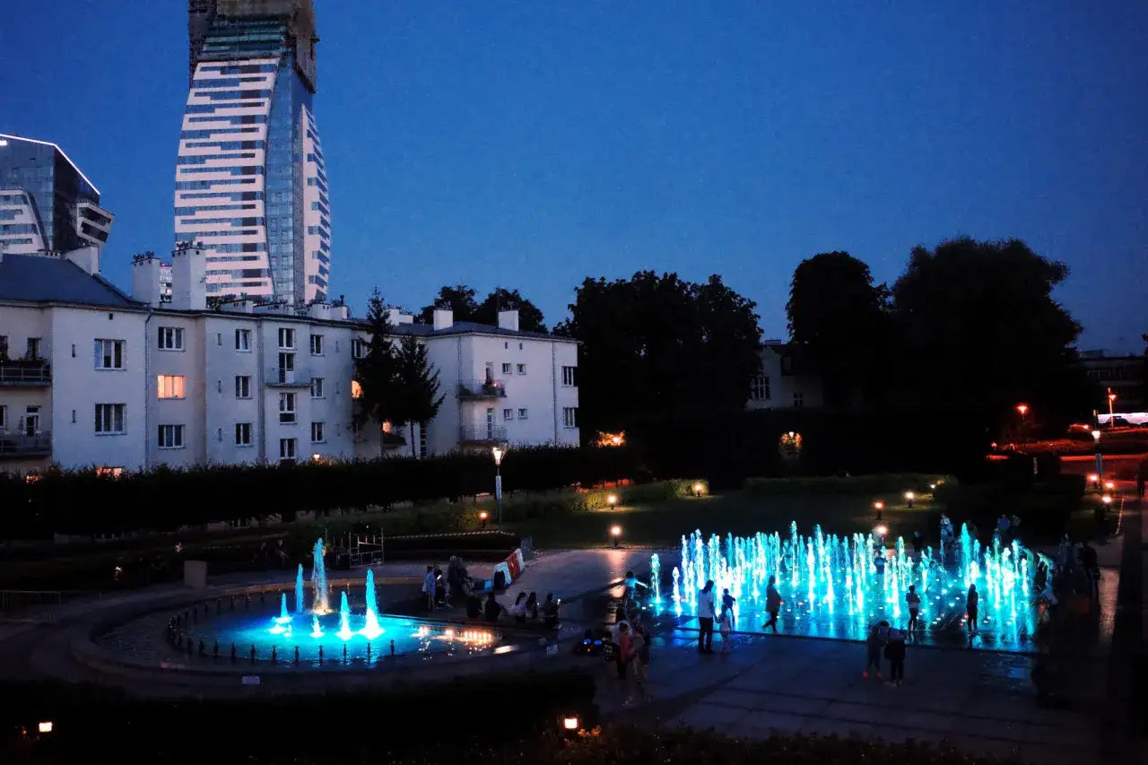 Rzeszów Rynek nocą: rozświetlone fontanny i ludzie spacerujący po placu, w tle budynki mieszkalne i nowoczesny wieżowiec.